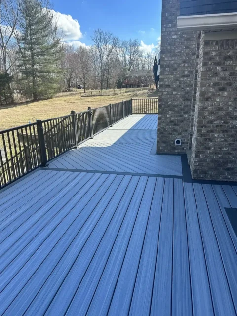 An outdoor deck constructed with gray or bluish-gray composite decking material. The deck is attached to a building with a lower section covered in dark blue siding and an upper section finished with reddish-brown brick. The deck features a black metal railing.