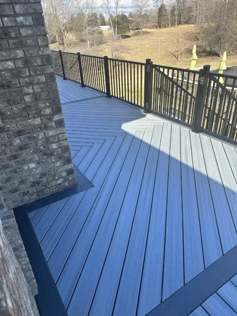 A close-up, elevated view of a gray composite deck. The deck features a pattern in which the boards meet at an angle in the middle, creating a V-shape that points away from the viewer. The deck has a black metal railing on the left side.
