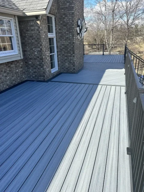 An elevated view of a gray composite deck attached to a brick house. The deck planks are laid horizontally and feature a fine wood-grain texture. The house has a dark gray-brown brick exterior, with white-trimmed windows visible on the lower level. A black metal railing lines the edge of the deck.